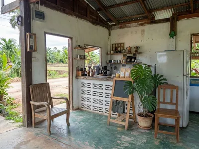 Simple cafe counter built from white breeze blocks with shelves of coffee gear, a monstera plant and wooden chairs opening onto a dirt lane