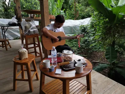 Man playing guitar beside a table of coffee pots and bagged green beans on a wooden deck