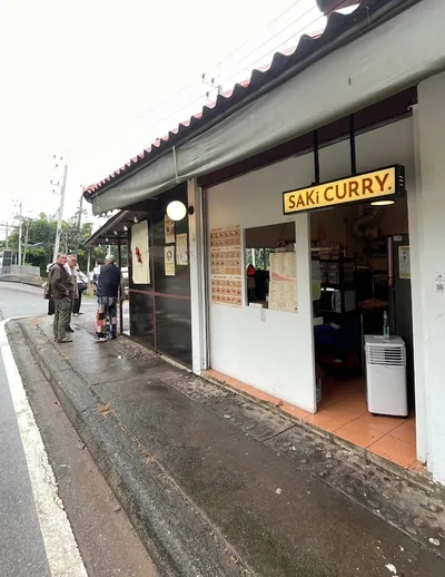 People waiting on a wet pavement at the counter window under a yellow Saki Curry sign