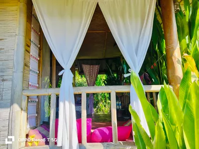 Bamboo hut with white curtains and pink floor cushions among banana leaves