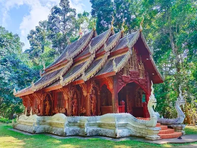 Carved red teak temple with tiered roofs and white naga balustrades under tall trees
