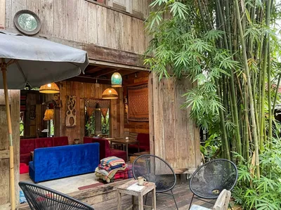 Open timber cafe front with a blue velvet sofa and red seats inside, string chairs on the patio beside a bamboo grove